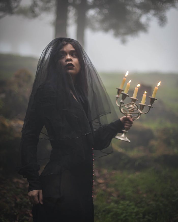 pexels photo 34489229 A mysterious woman in black attire holds a candelabra in a foggy forest setting.